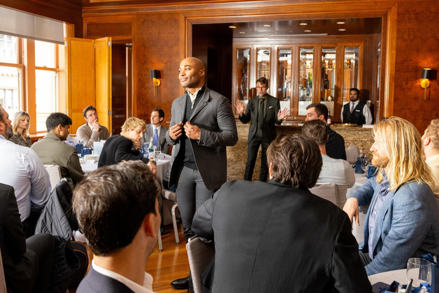A man in a suit stands speaking in front of a seated audience at a formal indoor event with wood-paneled walls and a bar in the background.